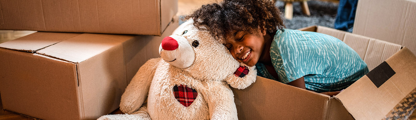 IMAGE: Young girl in moving box smiling and leaning head on large teddy bear.