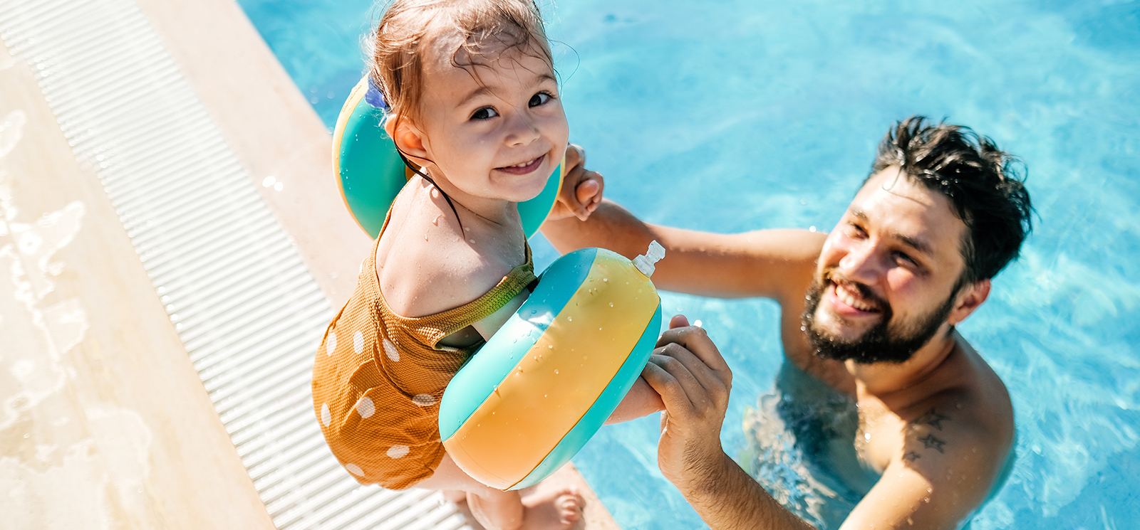 IMAGE: Man in pool holding young daughter's hand who is standing at the edge looking back at the camera