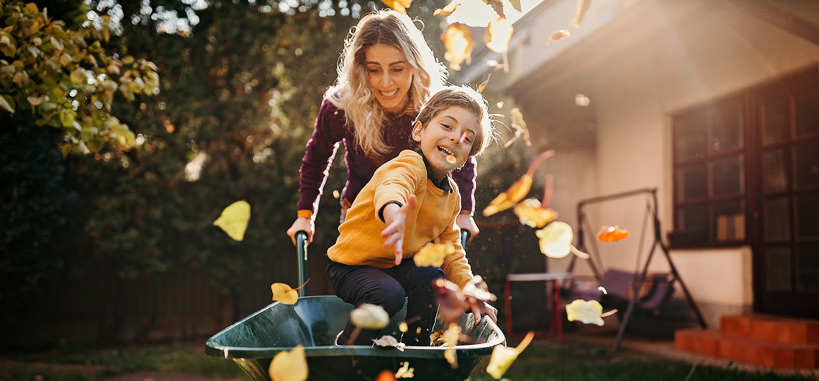 IMAGE: mom pushing young son in wheelbarrow with leaves falling.