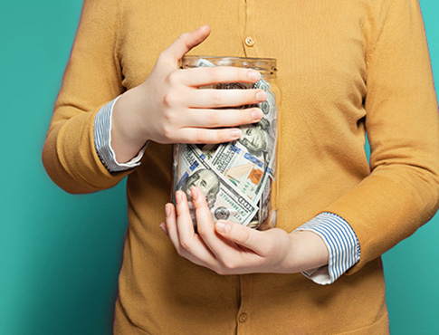 IMAGE: close cropped image of woman in yellow sweater holding jar of cash.